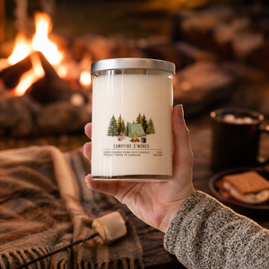Hand holding a candle labeled 'Campfire S'mores' in front of a cozy fireplace.