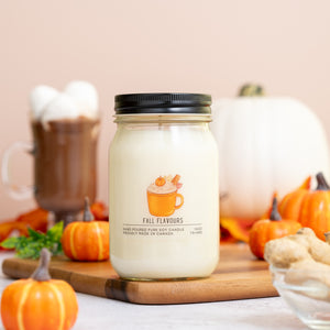 Candle labeled 'Fall Flavours' on a wooden board with pumpkins and hot chocolate in the background.