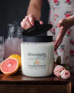 Serendipity candle with pink grapefruit and rosemary fragrance on a wooden surface with a person and fruit in the background.