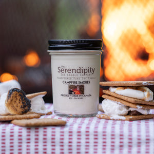Serendipity candle labeled 'Campfire Smores' with s'mores and graham crackers on a tablecloth in front of a campfire.