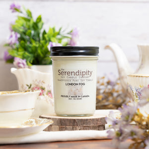 Serendipity candle labeled 'London Fog' on a wooden coaster with flowers and teacup in the background.