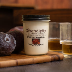 Serendipity candle with spiced bourbon plum scent on a wooden cutting board with plums and glasses in the background.